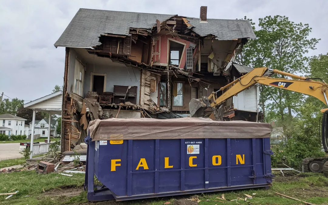 Excavator tearing down a house beside a purple Falcon Green Resources dumpster