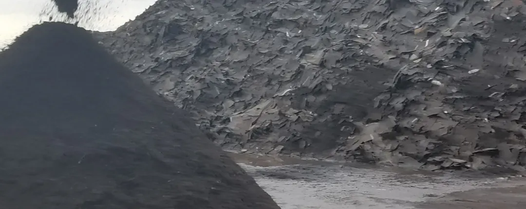Cono de material fino y oscuro de tejas asfálticas recicladas frente a una gran pila de tejas trituradas