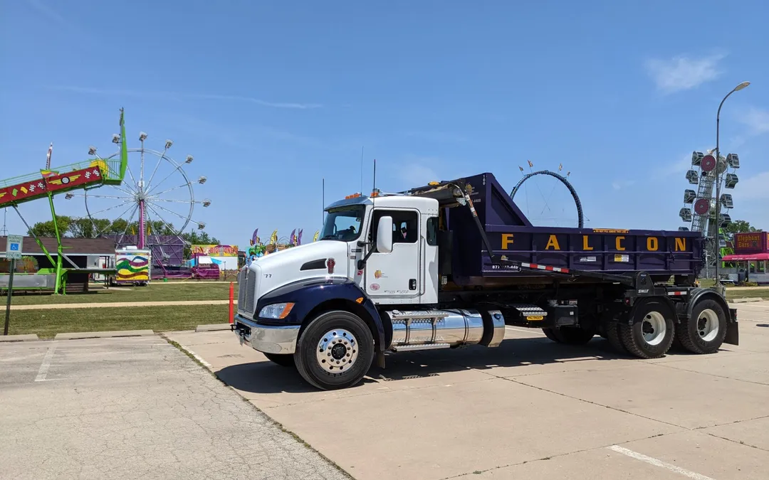 Falcon roll-off truck with a purple dumpster parked at a community fairground with carnival rides behind it