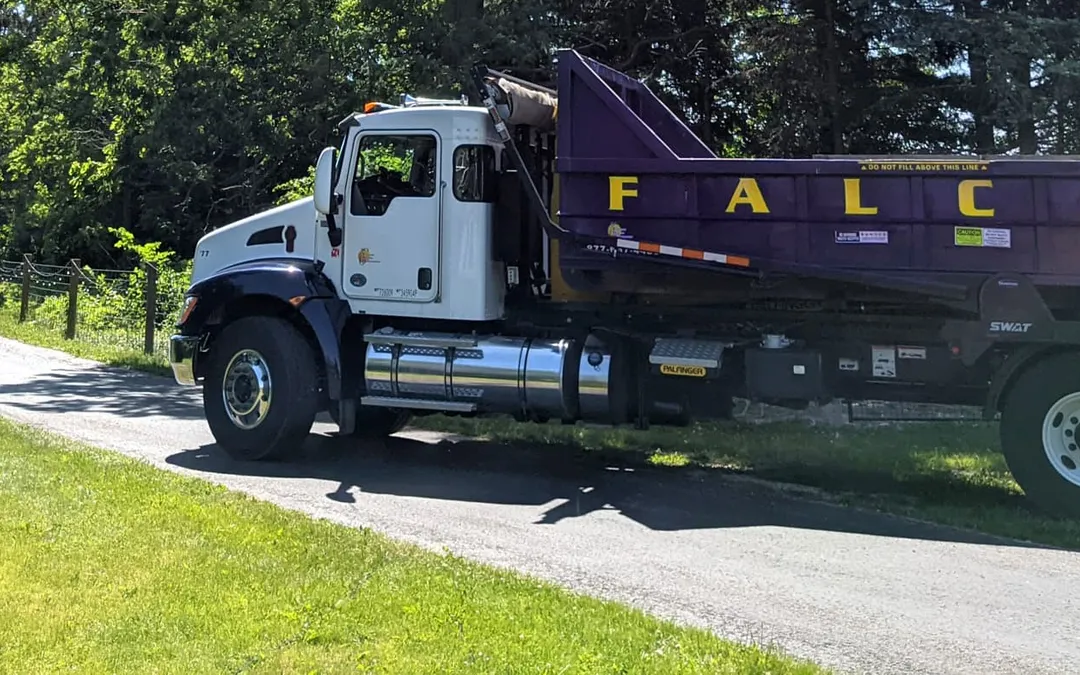 Falcon roll-off truck with a purple dumpster driving along a tree-lined country road