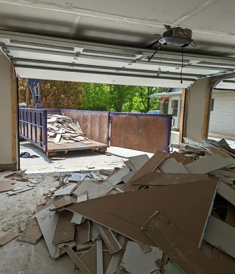 Drywall scraps covering a garage floor with a purple roll-off dumpster waiting in the driveway